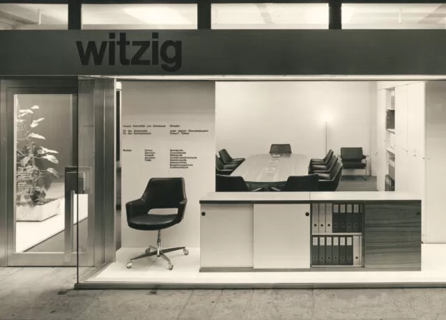 Historisches Schwarz-Weiss-Foto eines Büros mit dem Schriftzug "witzig" über der Eingangstür. Ein Konferenzraum mit einem grossen Tisch und schwarzen Stühlen ist durch das Fenster sichtbar. Vorne ist ein schwarzer Bürostuhl ausgestellt.