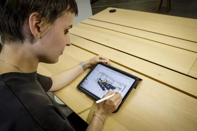 Eine Frau zeichnet auf einem Tablet, während sie an einem Holztisch sitzt. Der Raum hat eine minimalistische Einrichtung mit hellen Holzmöbeln und einem modernen Ambiente.