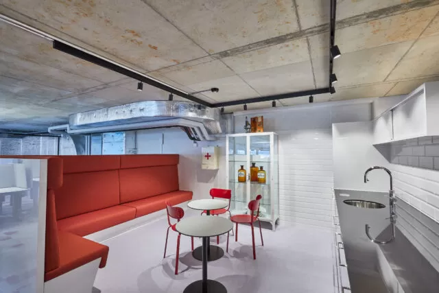 Moderner Pausenraum mit rotem Sofa, kleinem Tisch und Stühlen, Küchenzeile, industriell anmutende Decke mit sichtbaren Rohren, heller Bodenbelag. Requisiten aus dem Pharma und Gesundheitsbereich im Hintergrund.